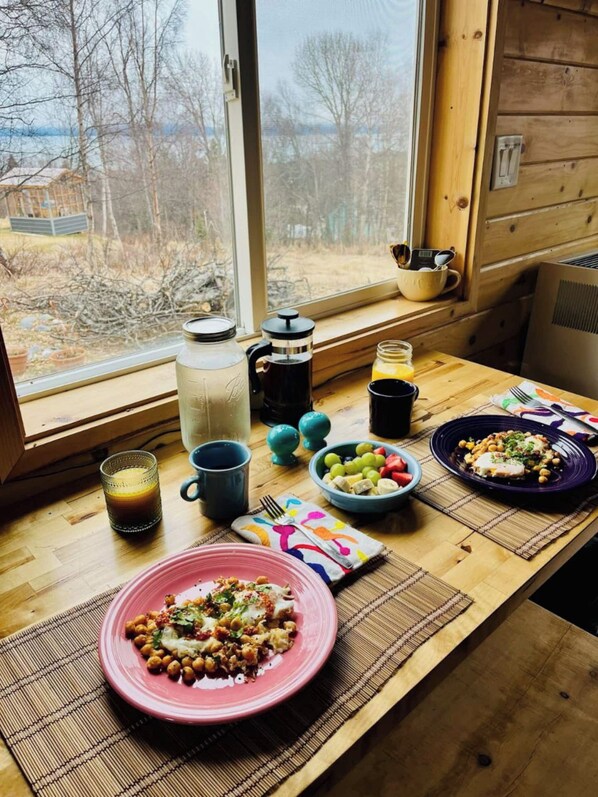 A breakfast table with a variety of food items, a coffee maker, and a view of the outdoors through a window.