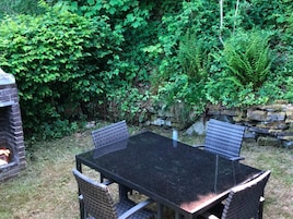 Un patio exterior con una mesa de vidrio y sillas de mimbre, rodeado de exuberante follaje y un muro de piedra.