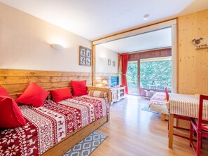 A cozy room with a wooden bed, red pillows, a small table with a lamp, and a view of the outdoors through a large window. - Les Deux Alpes Ski Apartment (Les Deux Alpes)