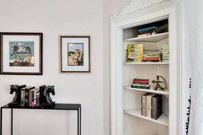 A room with a white bookshelf, framed pictures on the wall, and a black console table with books and decorative horse figures. - Chic 2 Bedroom Flat in Historic District - Wilmington (Wilmington)