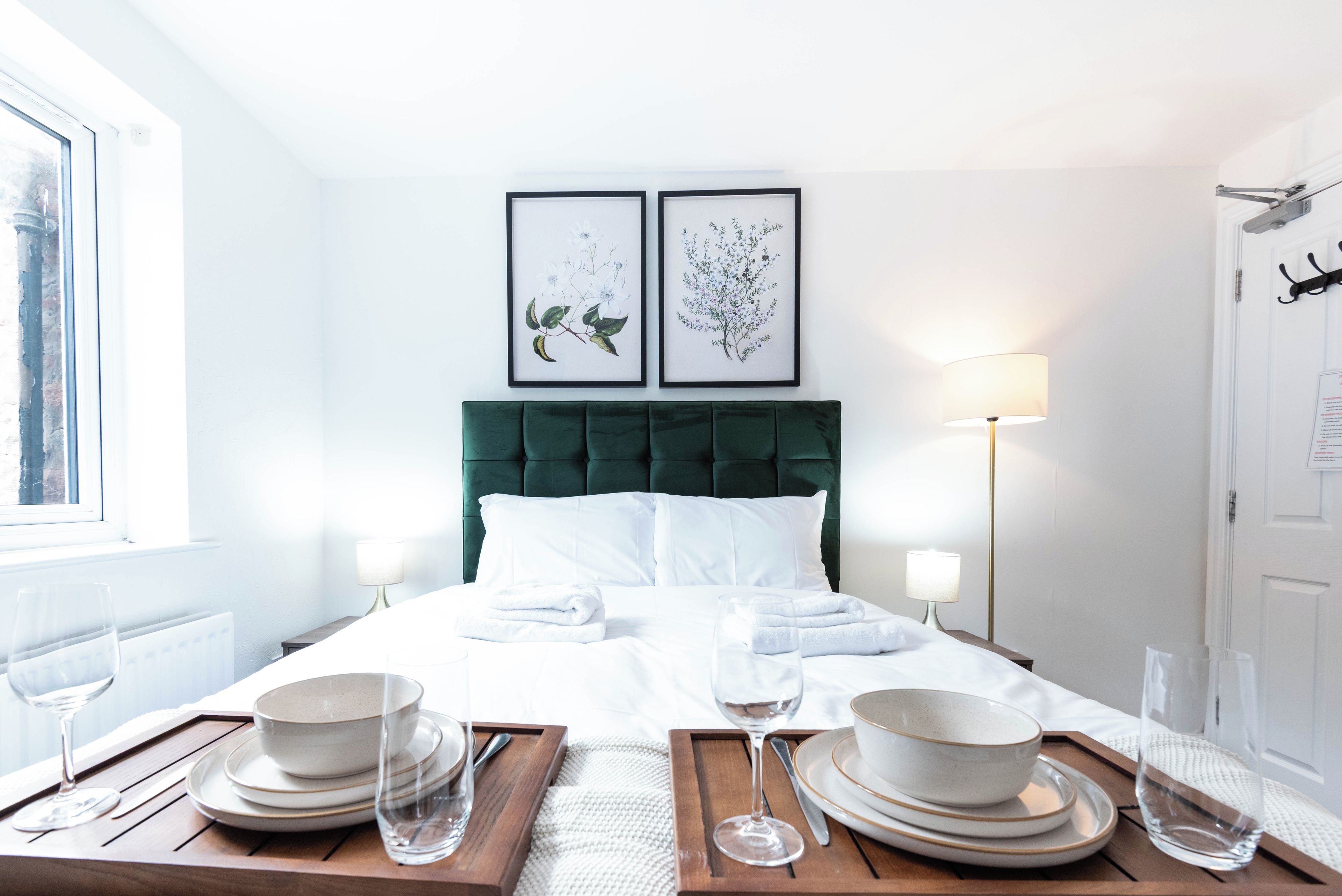 A hotel room with a bed, two framed floral artworks, bedside lamps, and a dining area with plates and glasses.