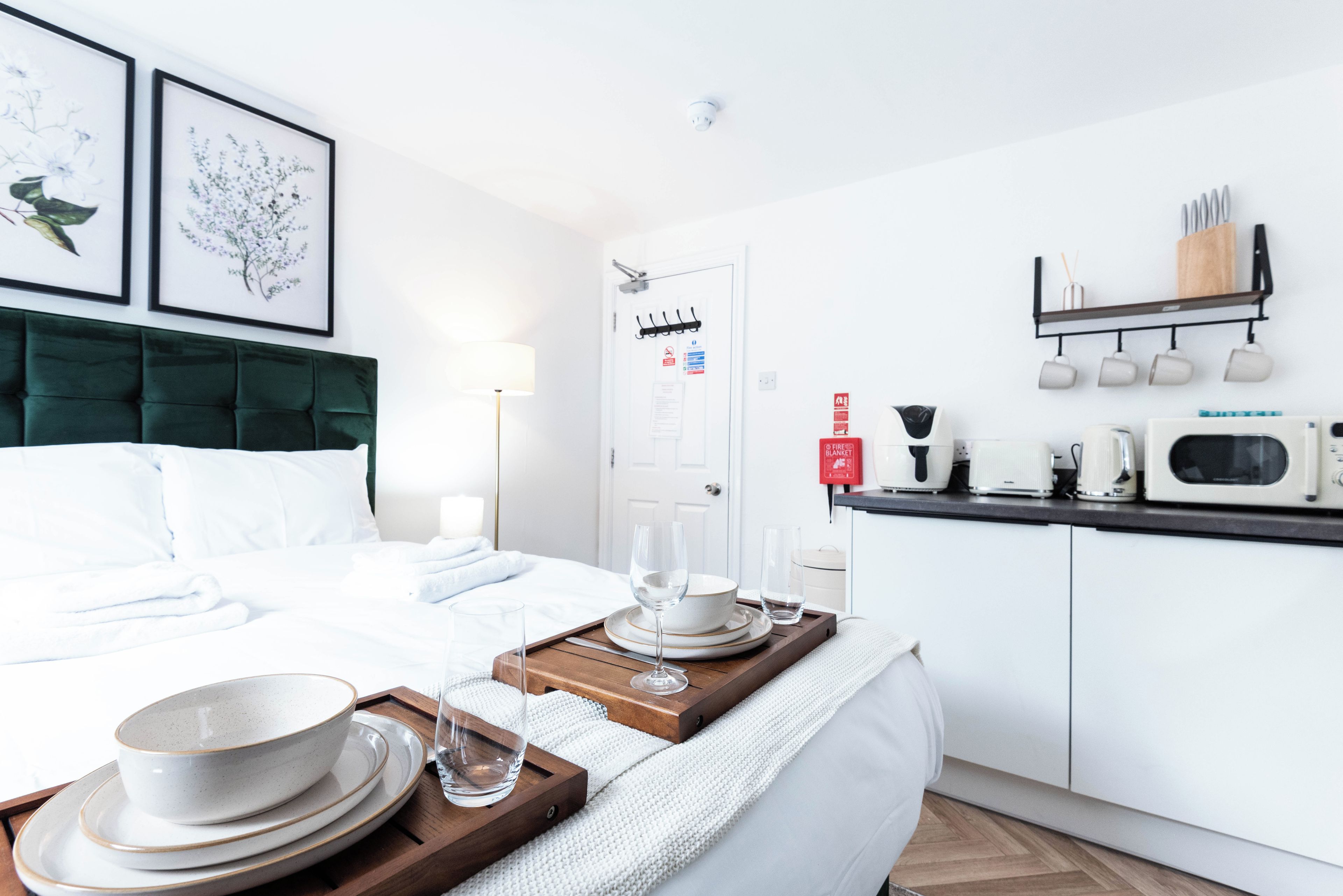 A hotel room with a bed, a bedside table with a tray of dinnerware, a kitchen area with a microwave, and a wall-mounted shelf with cups and a knife.