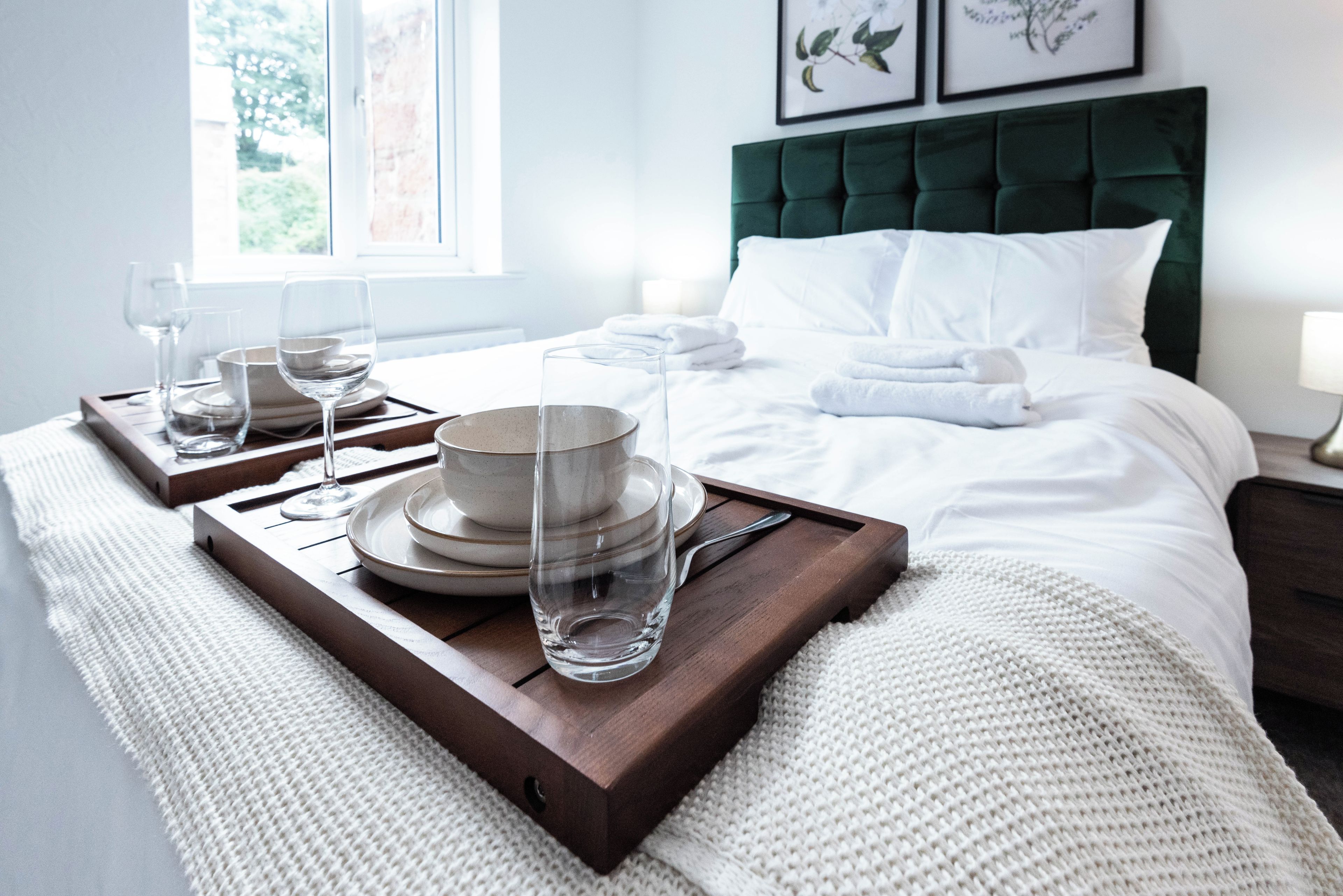 A neatly made bed with white linens, a green upholstered headboard, and a tray with dinnerware and glasses.