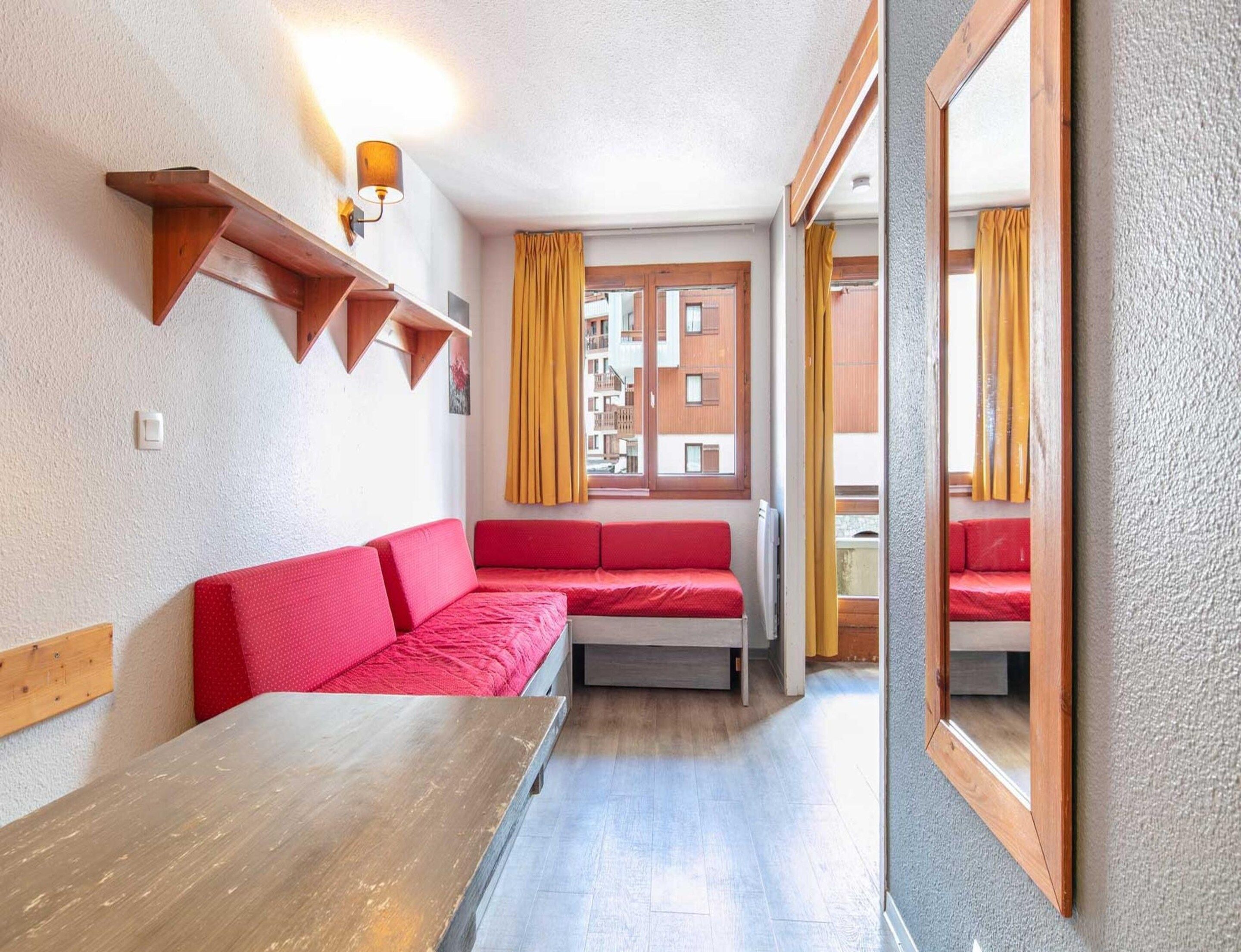 A compact room with a wooden table, red sofa, and a window with yellow curtains.