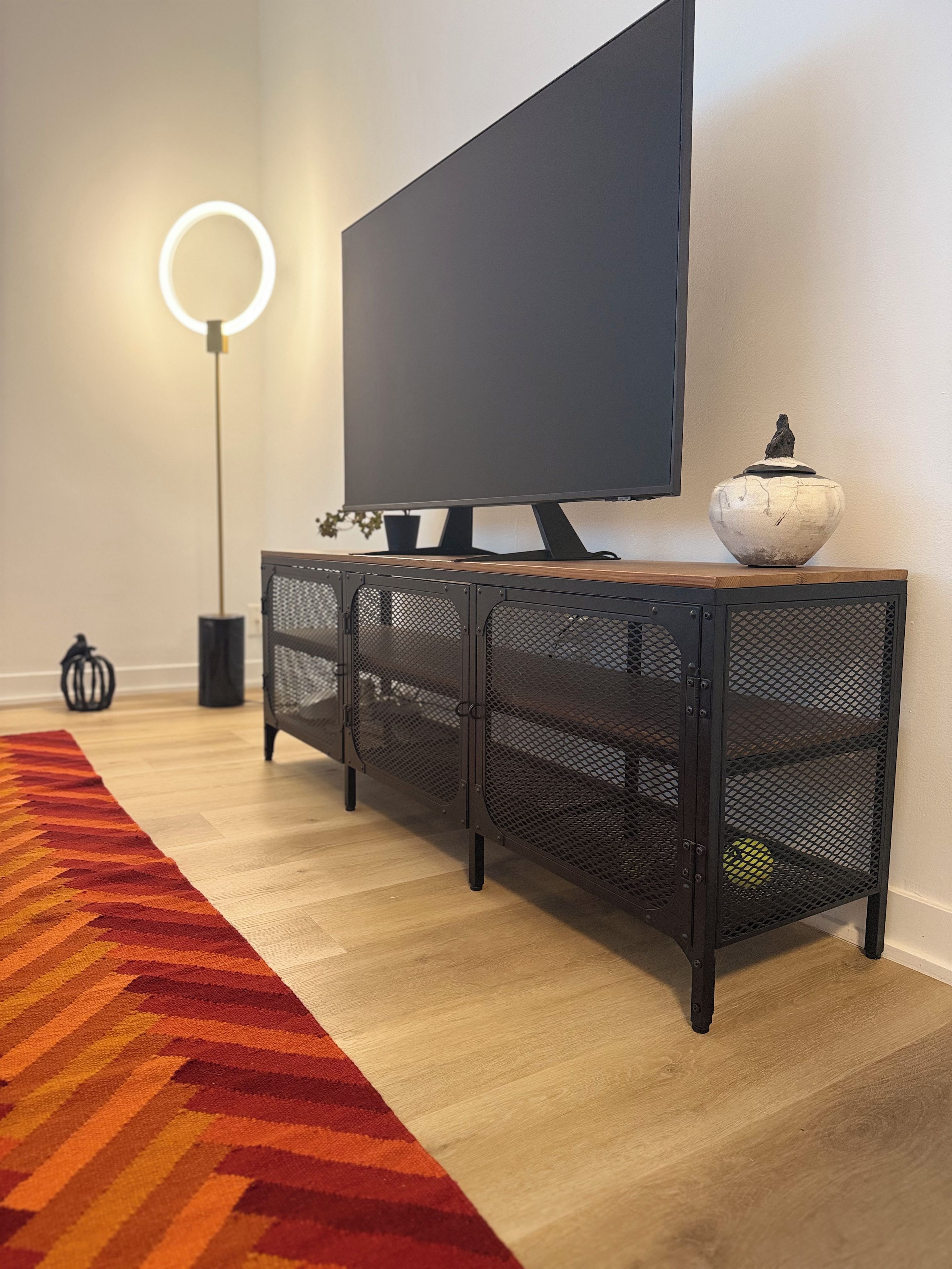 Un salon moderne avec un téléviseur à écran plat posé sur une console en métal noir, un lampadaire rond et un tapis à motifs.