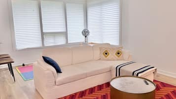 En stue med en beige sofa, et mønstret tæppe, et rundt sofabord og et vindue med skodder.