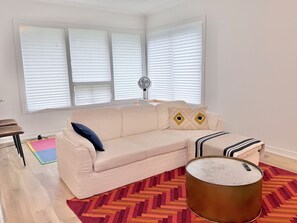 Ein Wohnzimmer mit einem beigefarbenen Sofa, einem gemusterten Teppich, einem runden Couchtisch und einem Fenster mit Jalousien.
