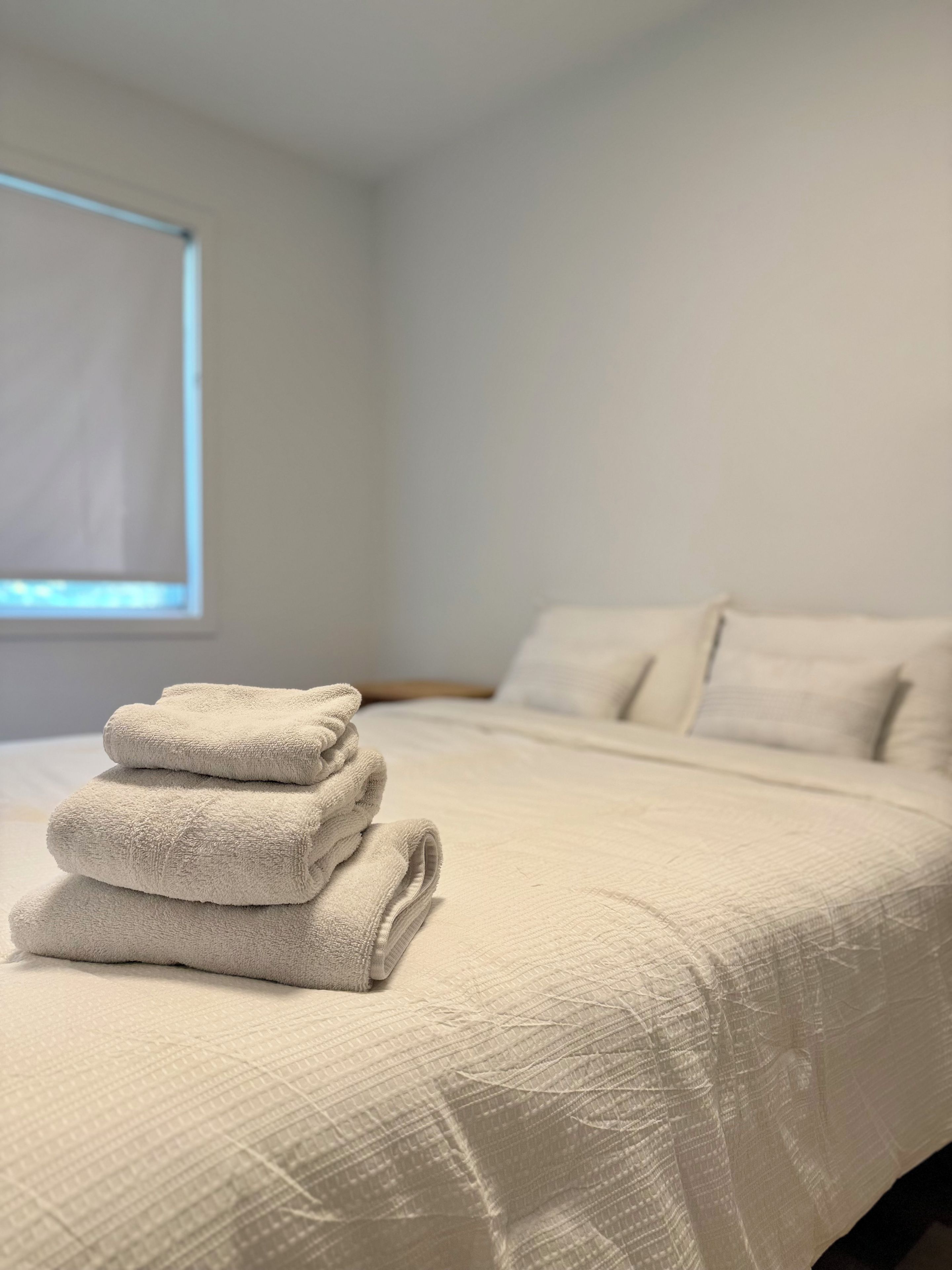 A neatly made bed with a folded white towel on top, a window with blinds, and a plain wall.