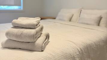 A neatly made bed with a folded white towel on top, a window with blinds, and a plain wall.