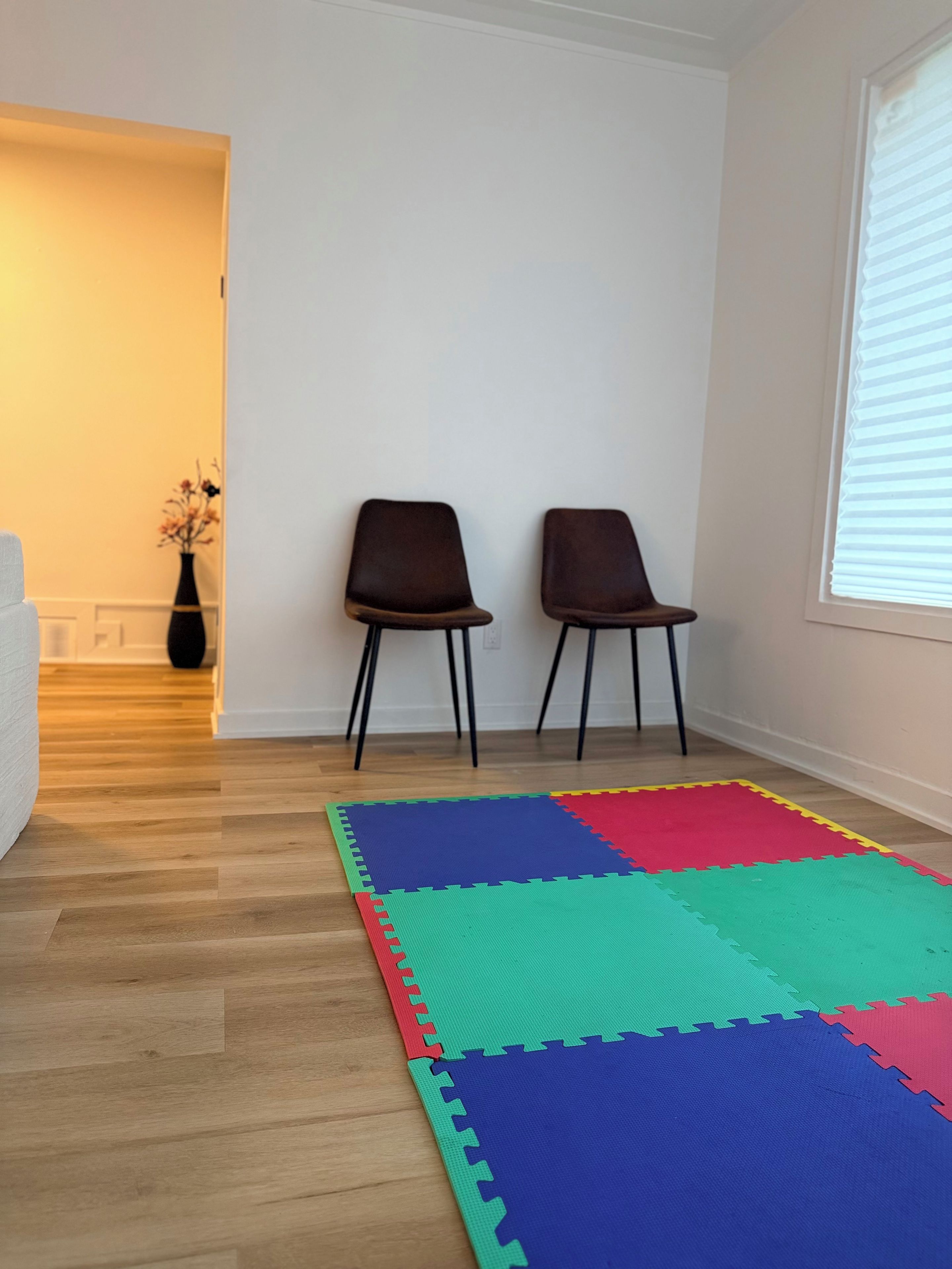 Ein Zimmer mit Holzboden, zwei Stühlen, einer bunten Schaumstoffmatte, einer Blumenvase und einem Fenster mit Jalousien.