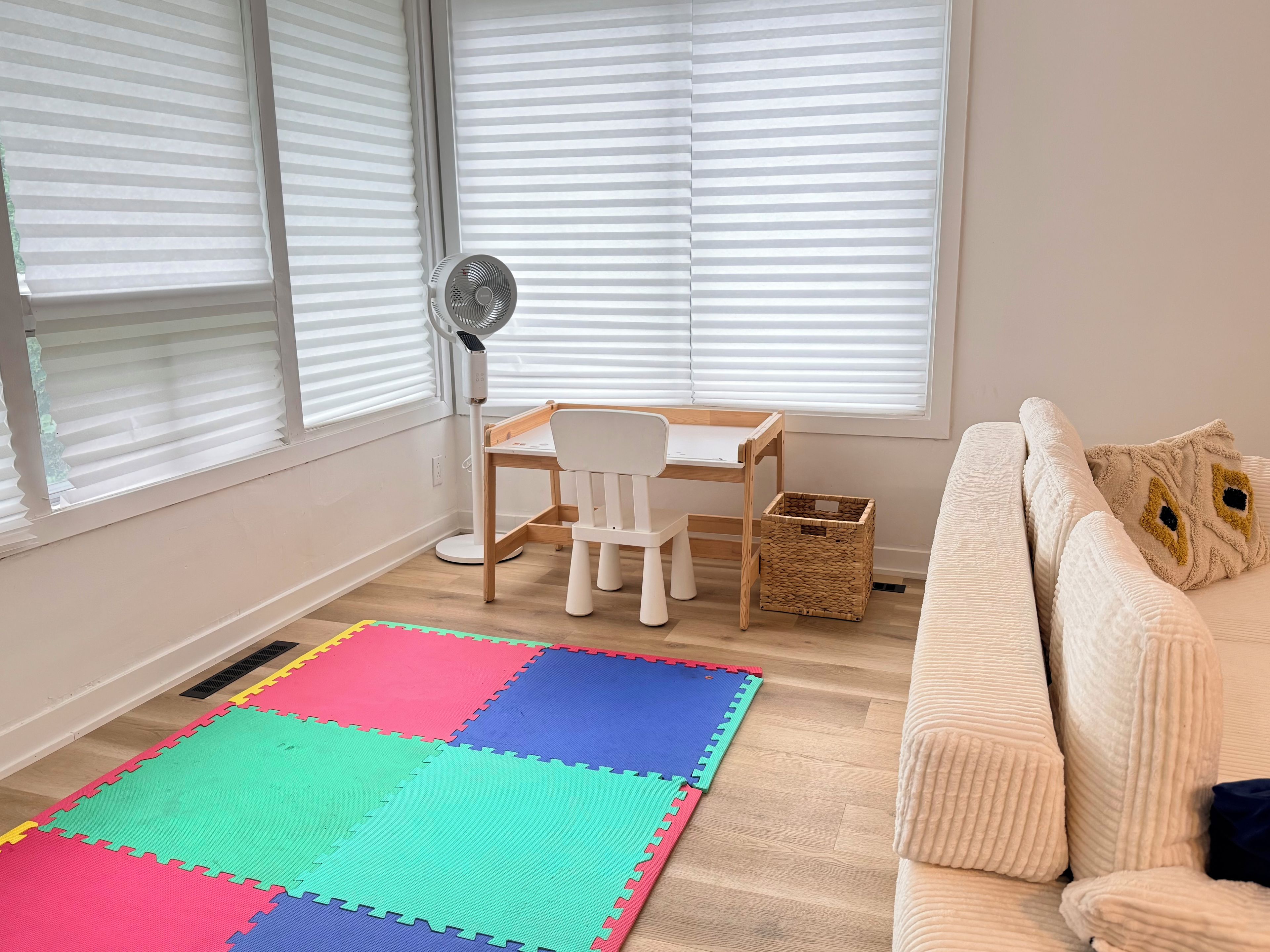 Una habitación con piso de madera, un tapete de espuma de colores, un escritorio blanco con una silla, un ventilador de pie y un sofá color beige con cojines decorativos.