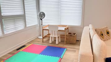 Una habitación con piso de madera, un tapete de espuma de colores, un escritorio blanco con una silla, un ventilador de pie y un sofá color beige con cojines decorativos.