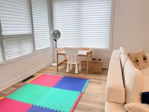A room with a wooden floor, a colorful interlocking foam mat, a white desk with a chair, a standing fan, and a beige sofa with decorative pillows. - La Maison Country Route 132 | Charmant Bungalow (Sainte-Catherine)