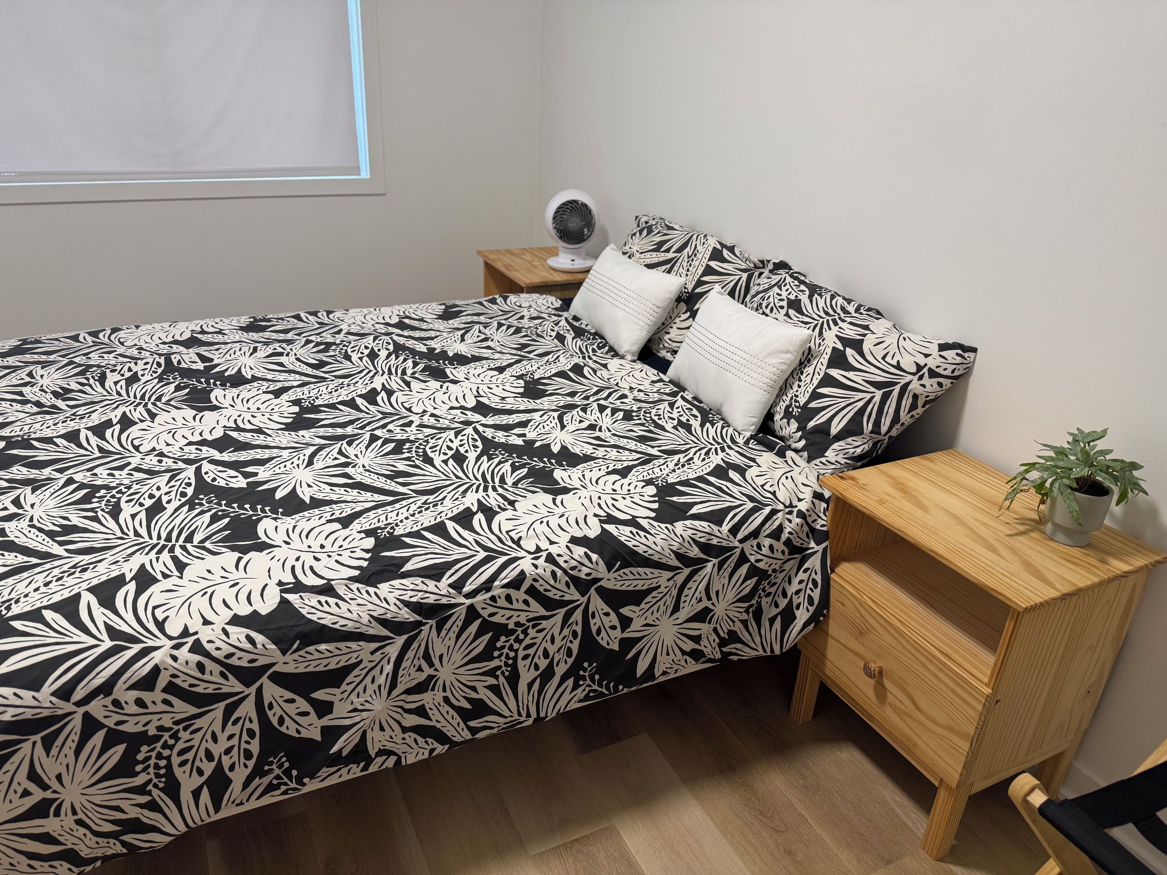 A bed with a black and white floral bedspread, a bedside table with a lamp, and a small potted plant on a wooden nightstand.