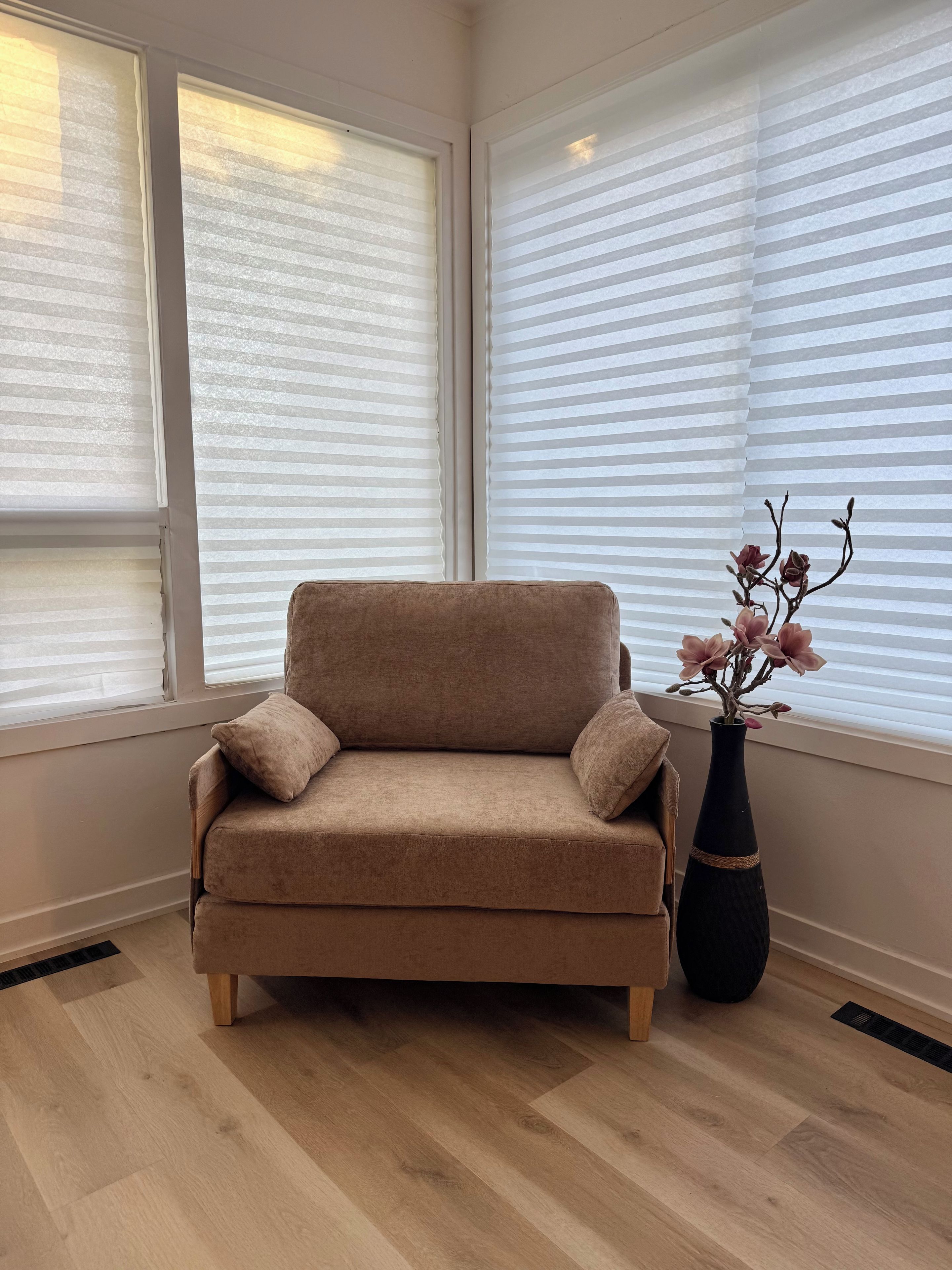 A beige sofa with cushions, a black vase with flowers, and a window with blinds.
