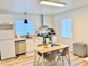 A modern kitchen with a dining area, equipped with a refrigerator, microwave, stove, and a small trash bin. - La Maison Country Route 132 | Charmant Bungalow (Sainte-Catherine)