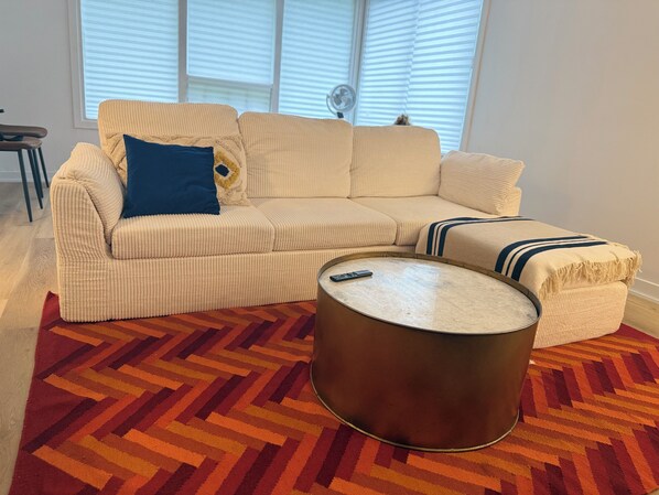 Un sofá color beige con un cojín azul, una manta a rayas y una mesa de centro redonda con un control remoto encima.