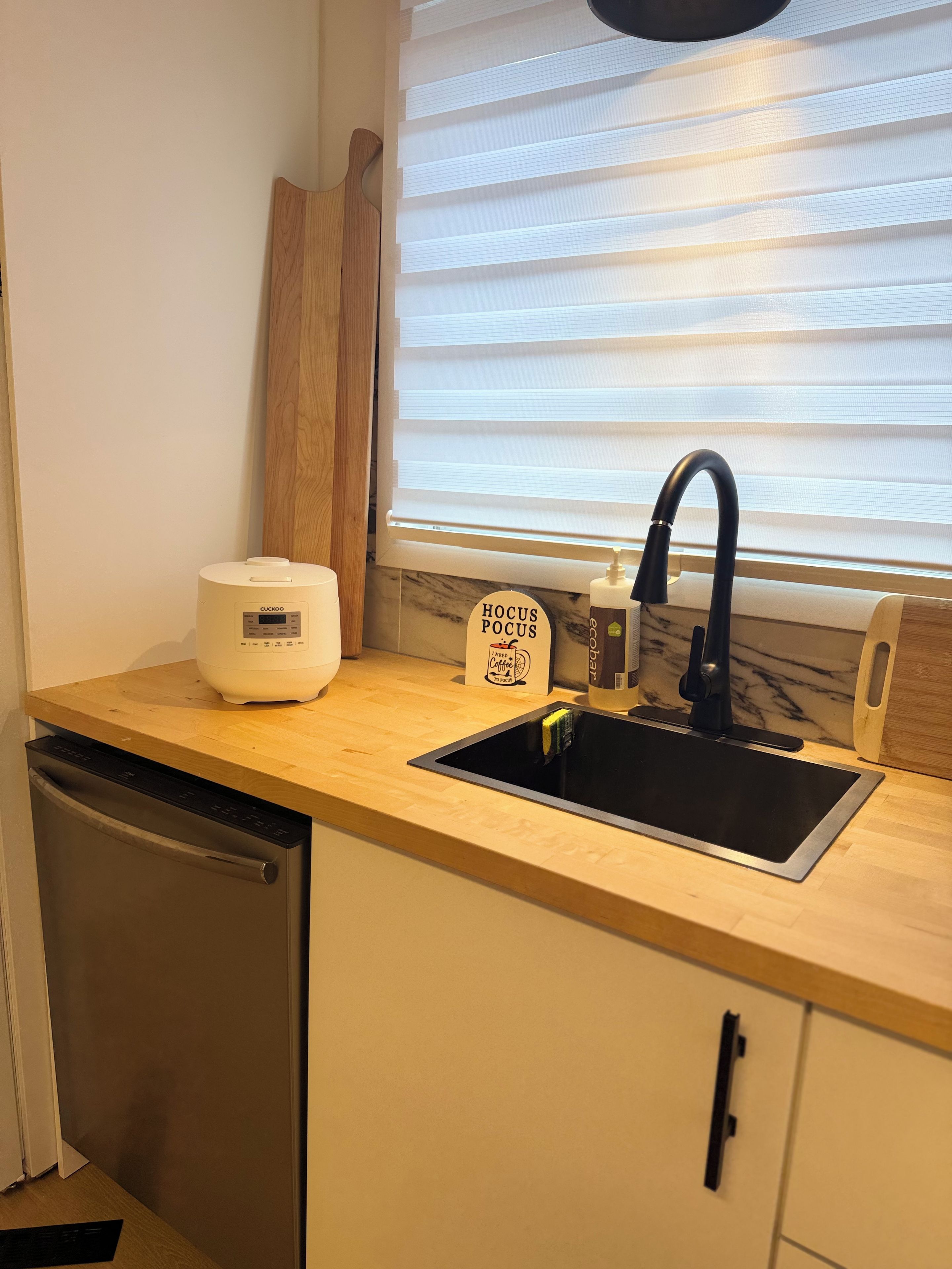 Eine Küche mit Geschirrspüler, Kaffeemaschine, Spüle und einem Fenster mit Jalousien.