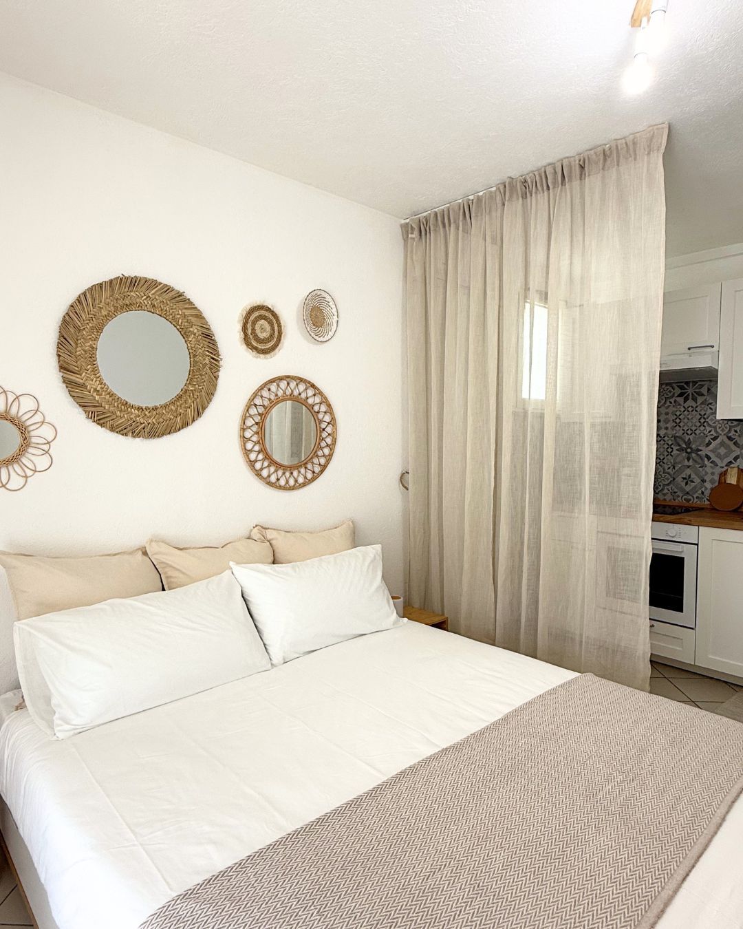 A bedroom with a bed, a window with sheer curtains, and a kitchenette in the background.