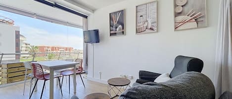 A modern living room with a sofa, a dining table with chairs, a TV mounted on the wall, and artwork.