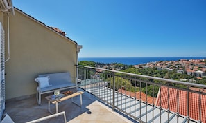 A balcony with a sofa, a table with a glass of wine, and a view of a coastal town. - Hedera Estate, Hedera A18 (Dubrovnik)