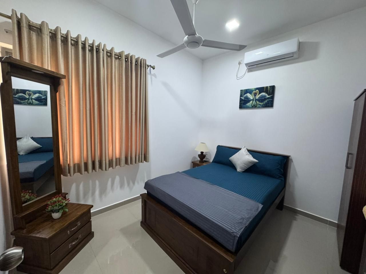 Een slaapkamer met een bed, een plafondventilator, een airconditioningapparaat, een nachtkastje met een lamp, een spiegel en gordijnen.