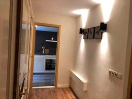 A hallway leading to a kitchen with a wooden floor, a white door, and a wall-mounted light fixture.