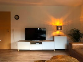 Una sala de estar con un televisor de pantalla plana sobre un mueble blanco, un reloj de pared y una lámpara de pie que proyecta un resplandor cálido.