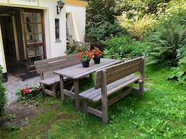 Un conjunto de banco y mesa de madera en un jardín, con un edificio blanco al fondo.
