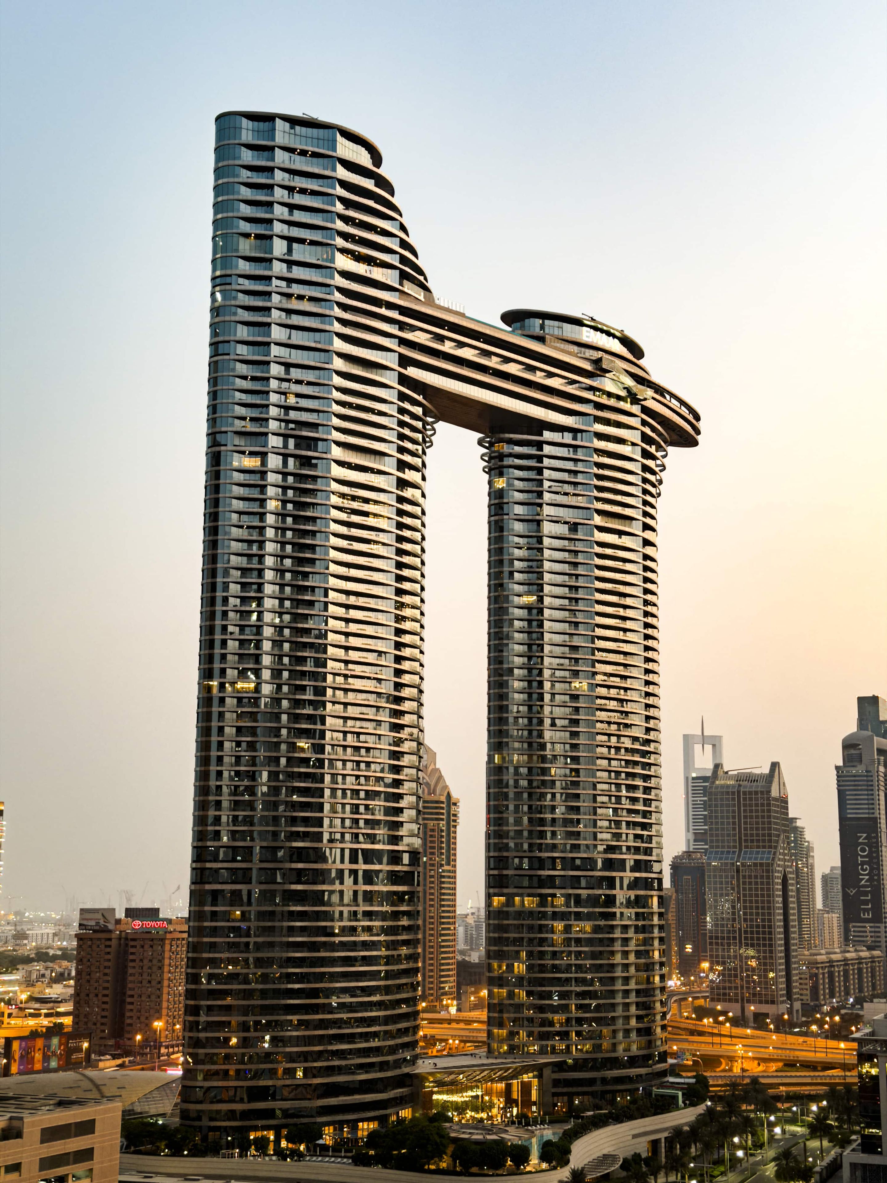 A modern high-rise building with a curved facade, multiple balconies, and a prominent overpass structure.