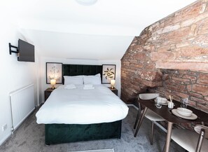 A hotel room with a large bed, a stone wall, a dining table with chairs, and a flat-screen TV mounted on the wall. - The Stanley Arms Hotel by Guestz (Seascale)