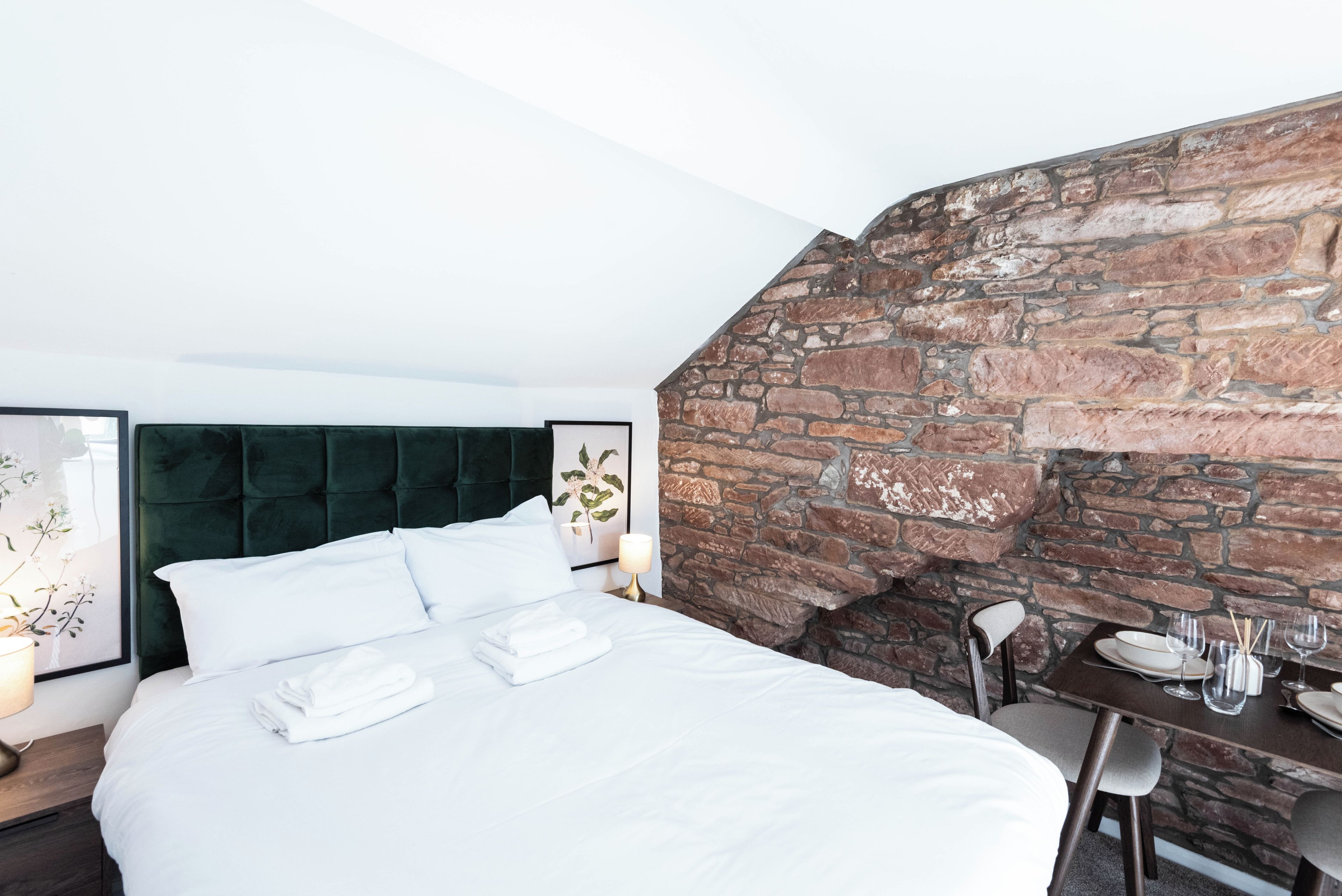 A hotel room with a bed, a headboard, a table with chairs, and a stone wall.