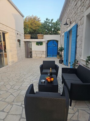 An outdoor seating area with wicker furniture, a table with a fruit bowl and a bottle, and a blue door in the background.