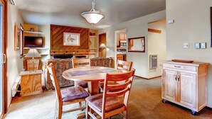 A dining area with a wooden table and chairs, a television, and a kitchen area with cabinets and a microwave. - Forest Condominiums (Keystone)