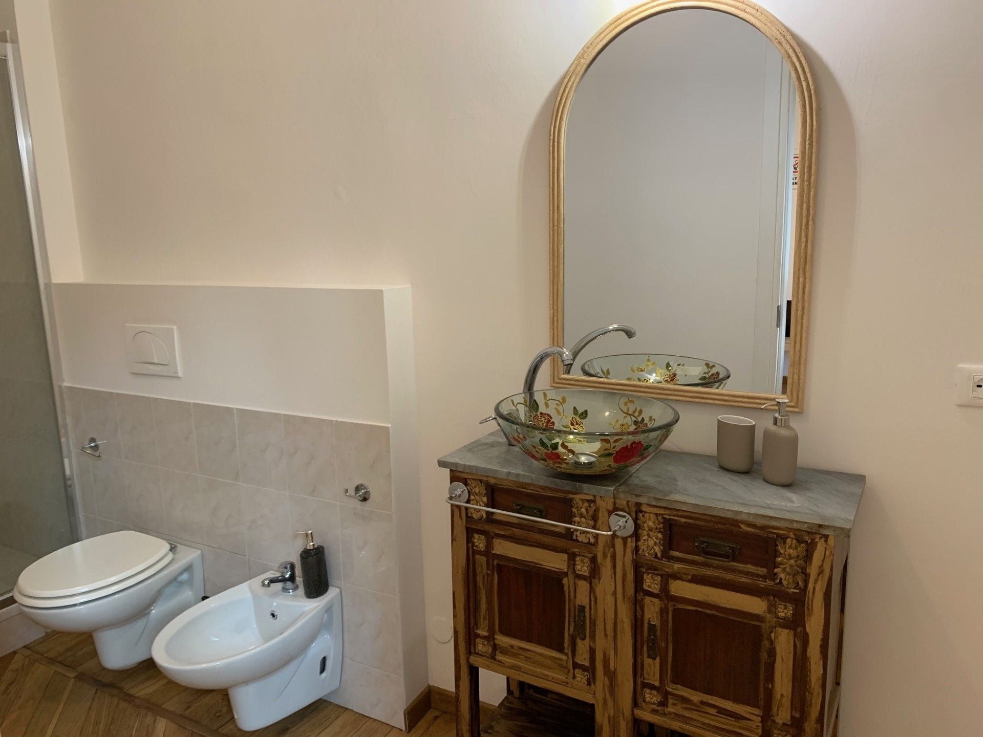 A bathroom with a toilet, bidet, vanity with a mirror, and a decorative glass bowl on the sink.