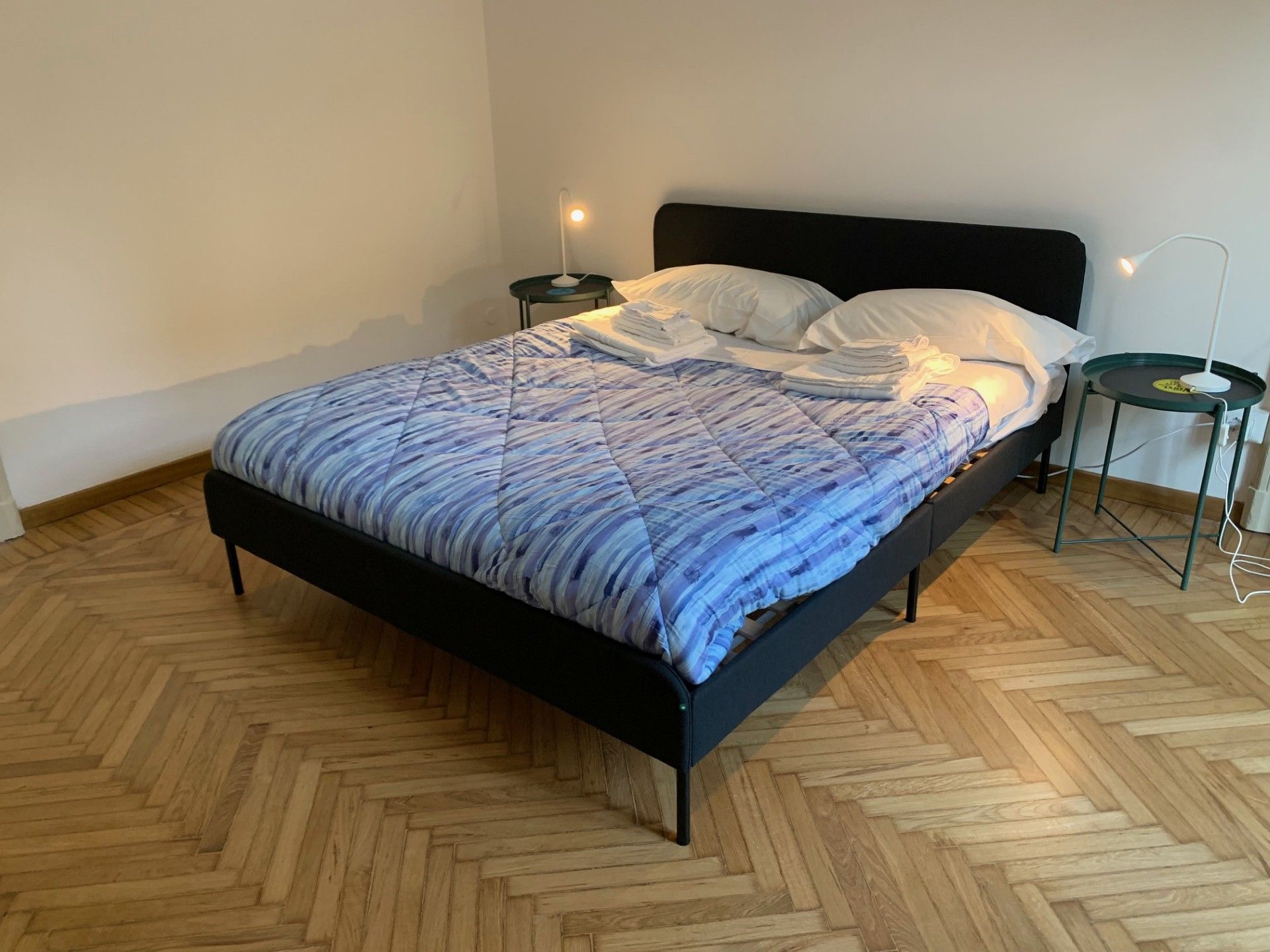 Una cama con una colcha estampada, dos mesitas de noche con lámparas y un suelo de madera con diseño de espiga.