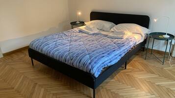 Una cama con una colcha estampada, dos mesitas de noche con lámparas y un suelo de madera con diseño de espiga.