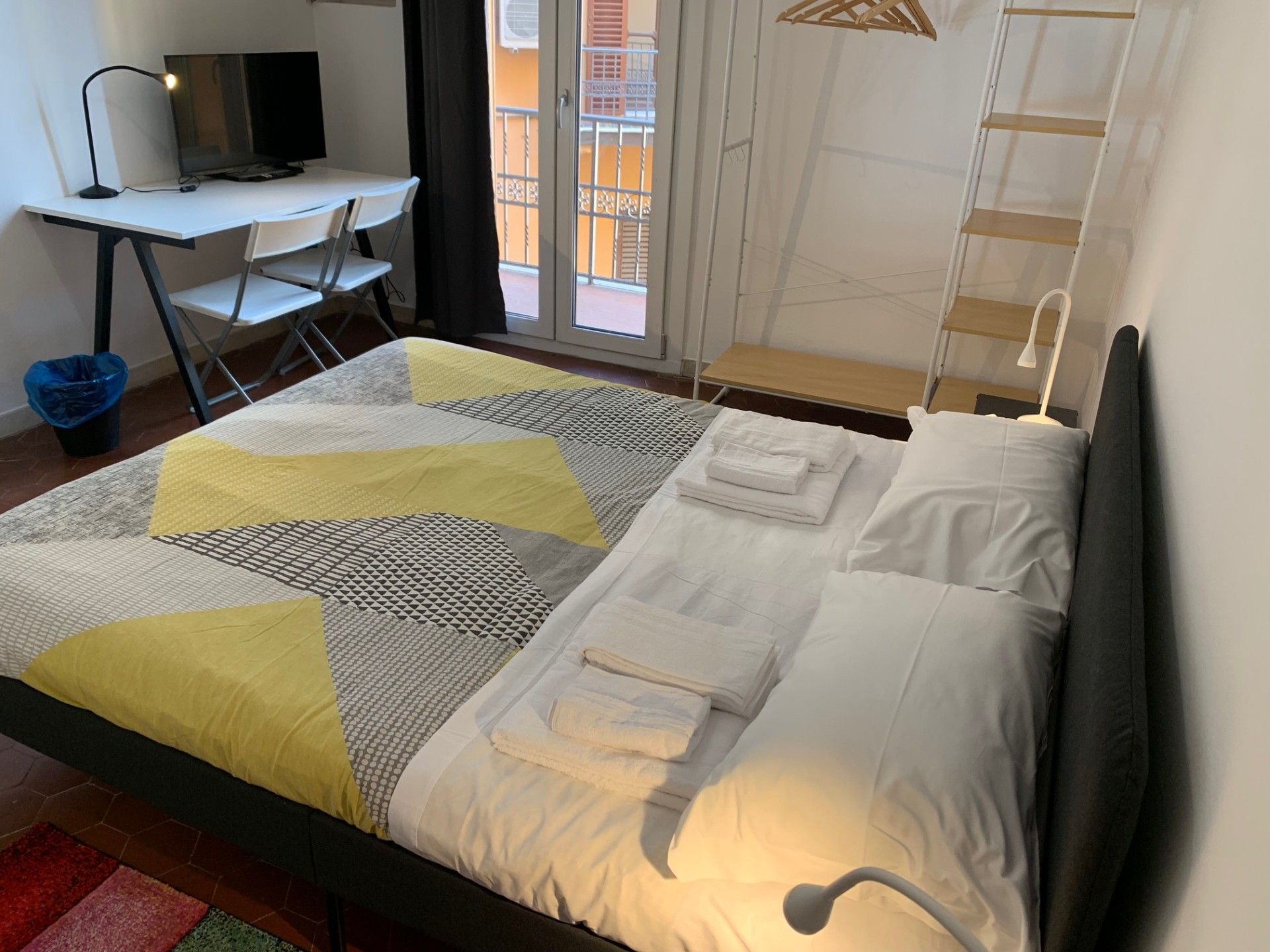 Ein ordentlich bezogenes Bett mit weißer und gelber Bettwäsche, ein Schreibtisch mit Computer und Stuhl sowie ein Fenster mit Blick auf Gebäude.
