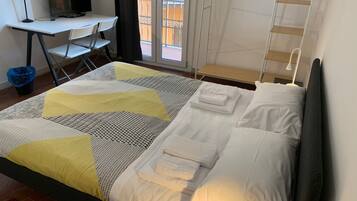 Ein ordentlich bezogenes Bett mit weißer und gelber Bettwäsche, ein Schreibtisch mit Computer und Stuhl sowie ein Fenster mit Blick auf Gebäude.