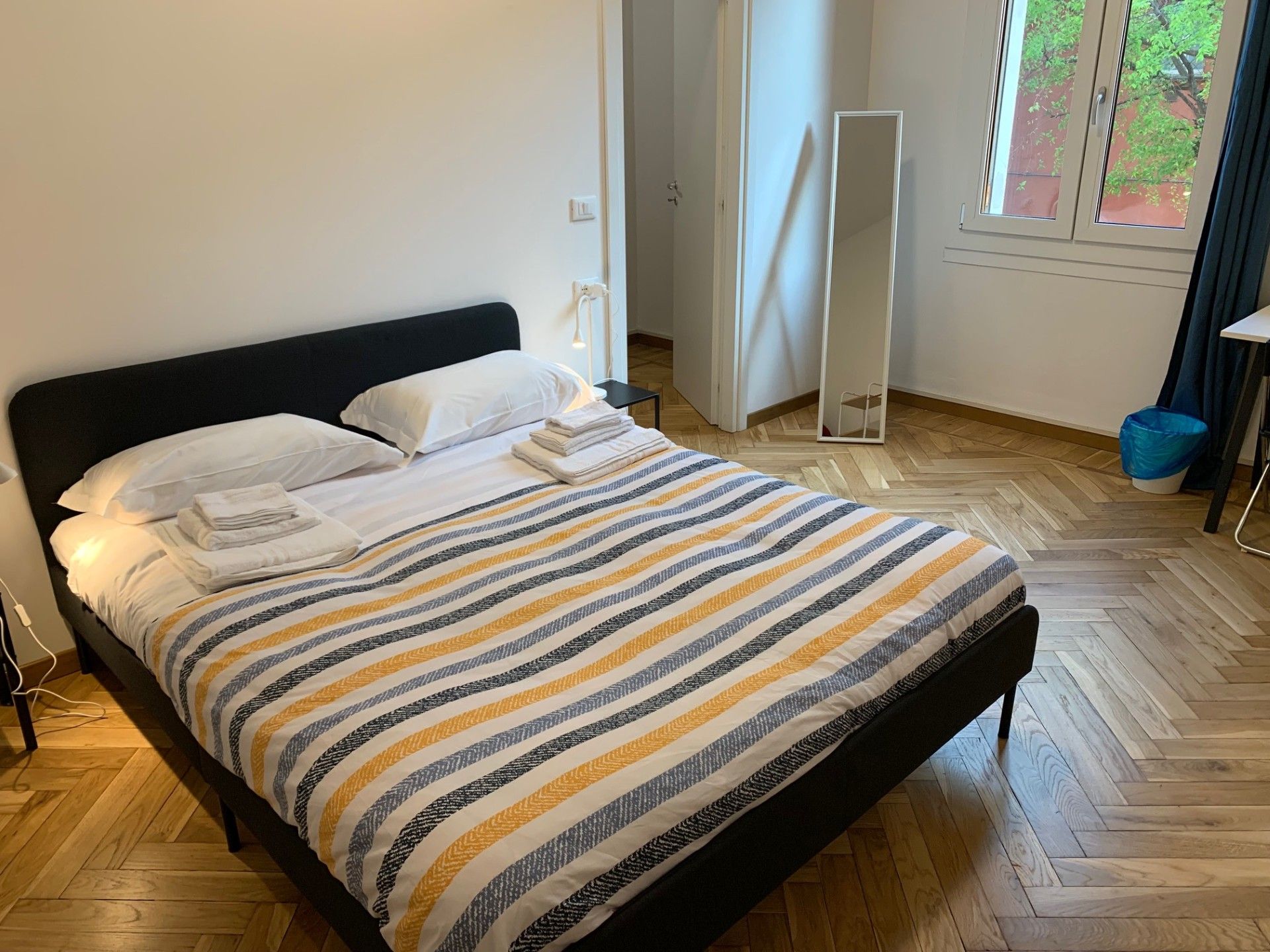 Ein ordentlich bezogenes Bett mit einem gestreiften Überwurf, ein Nachttisch mit Lampe und ein Fenster mit Vorhängen.