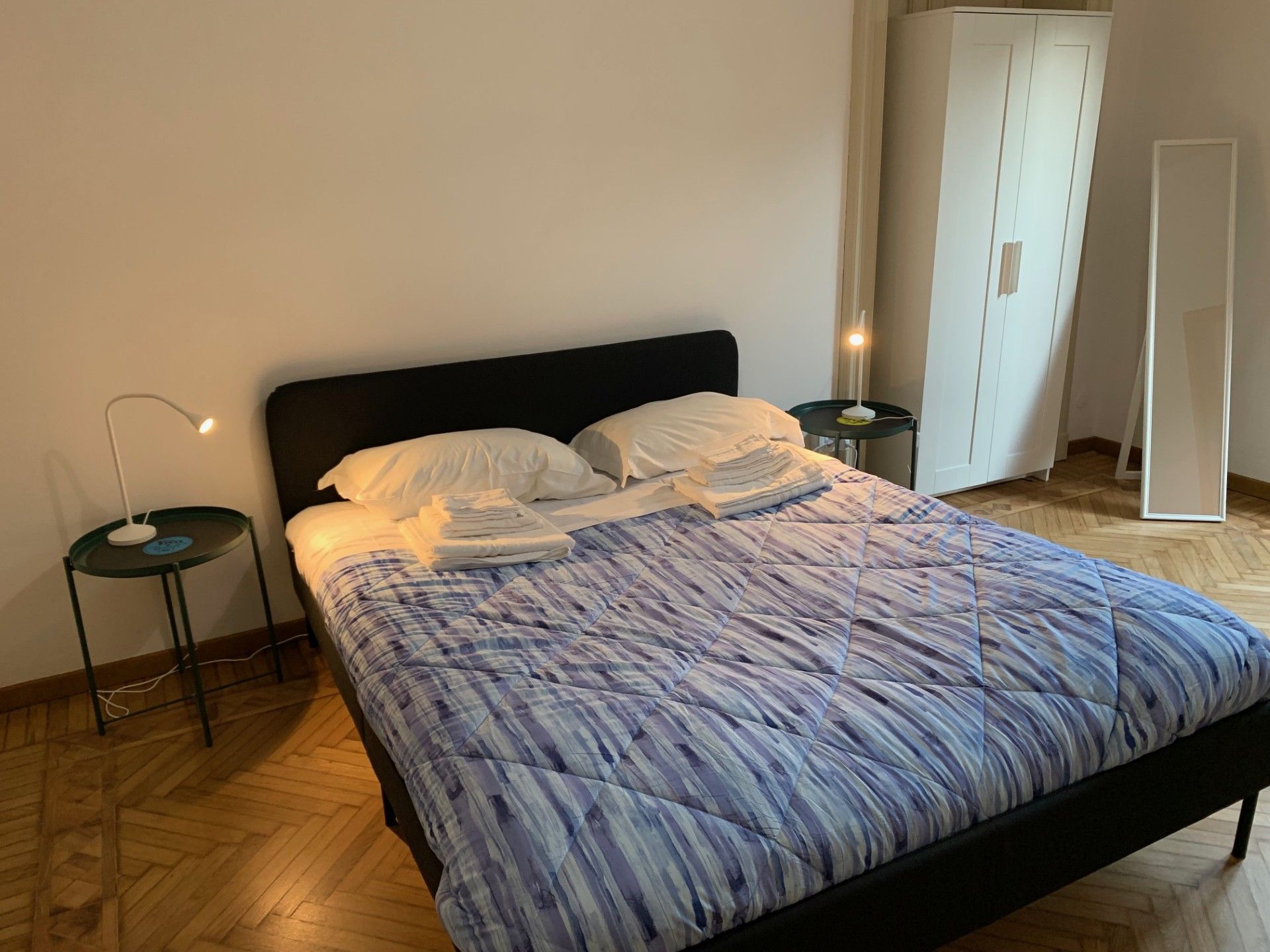 A bed with a blue and white patterned duvet, two bedside tables with lamps, and a wardrobe in the background.