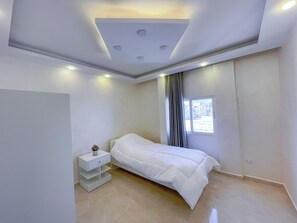 Een slaapkamer met een bed, een nachtkastje, een raam met gordijnen en inbouwspots.