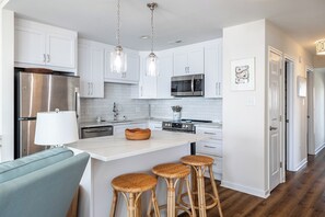 A modern kitchen with white cabinets, stainless steel appliances, a marble island, and wicker bar stools. - Ocean Reef 8-A2 | Oceanfront Condo (Emerald Isle)