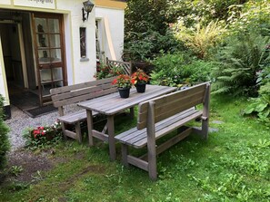 Un conjunto de banco y mesa de madera en un jardín, con un edificio blanco al fondo.