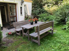 Un conjunto de banco y mesa de madera en un jardín, con un edificio blanco al fondo.
