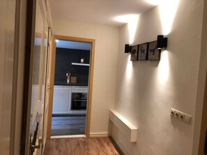 Un pasillo con suelo de madera, una puerta a la izquierda y una cocina visible a través de una puerta abierta.