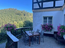 A balcony with a table and chairs, surrounded by flower boxes with pink flowers, overlooking a forested hillside.