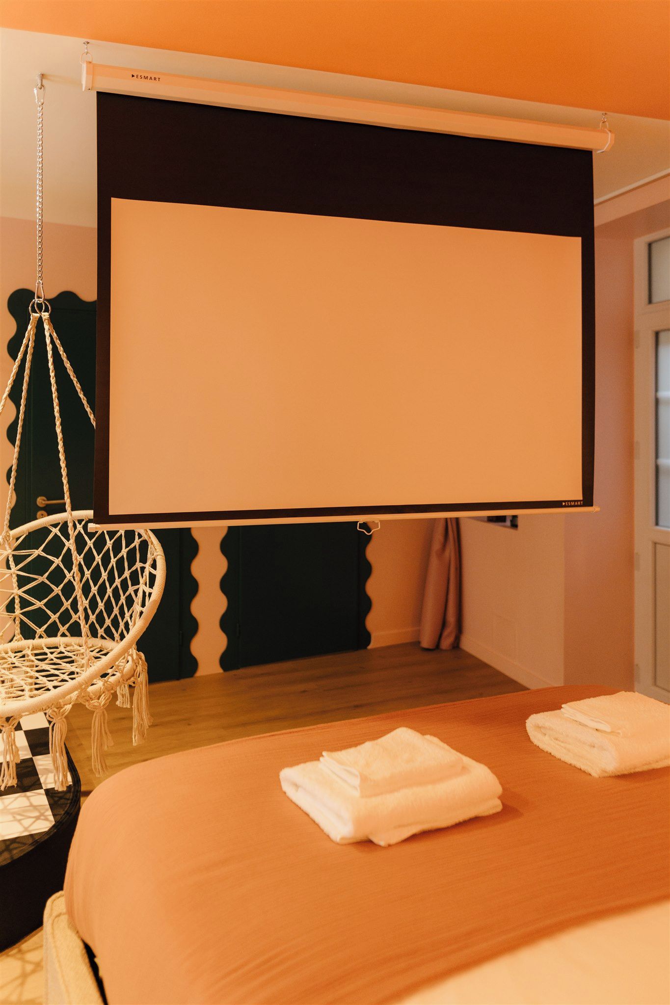A room with a bed, a macrame chair, a projector screen, and a window.