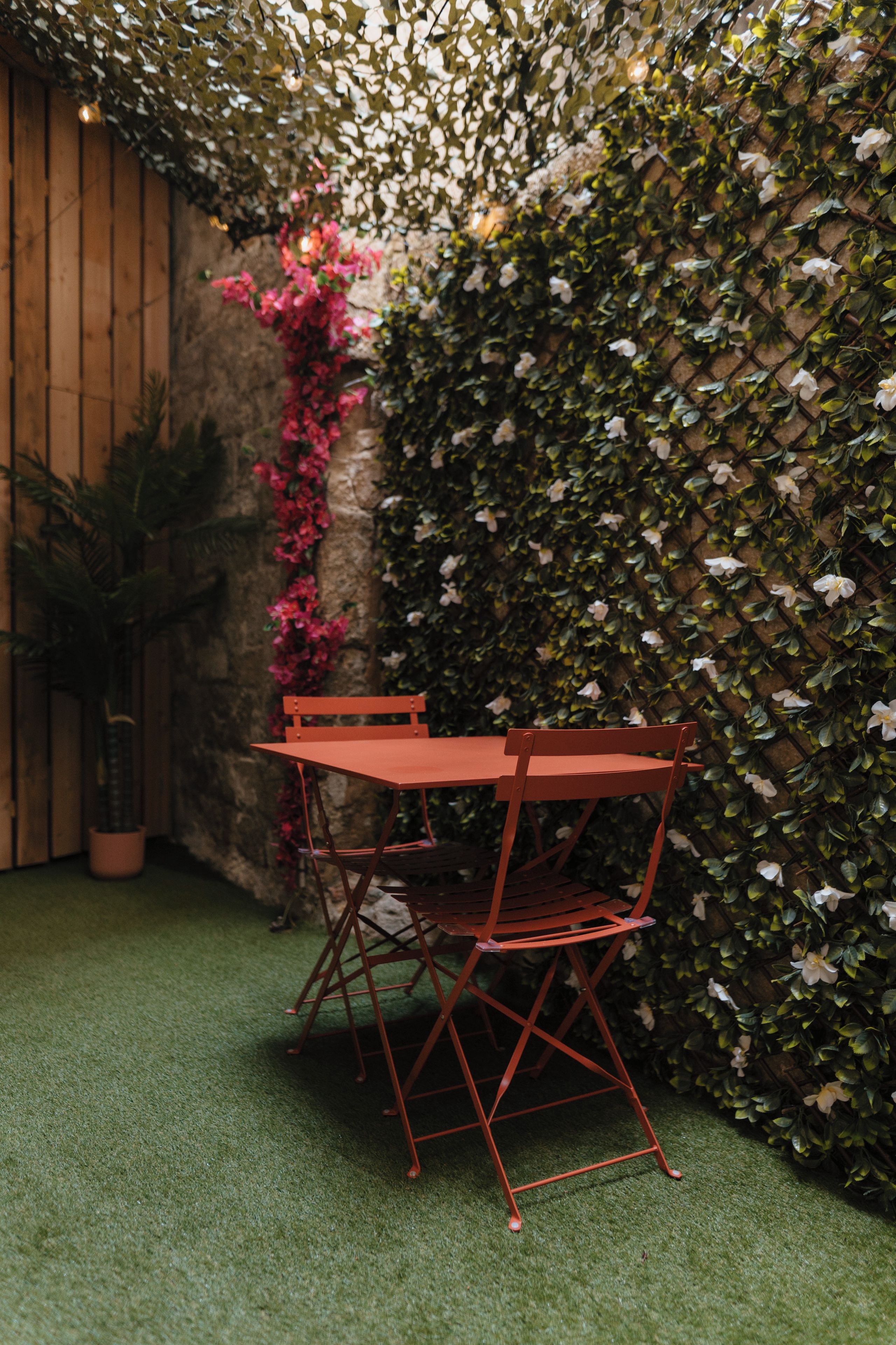 A red metal table and chair set against a wall covered in green foliage and pink flowers.