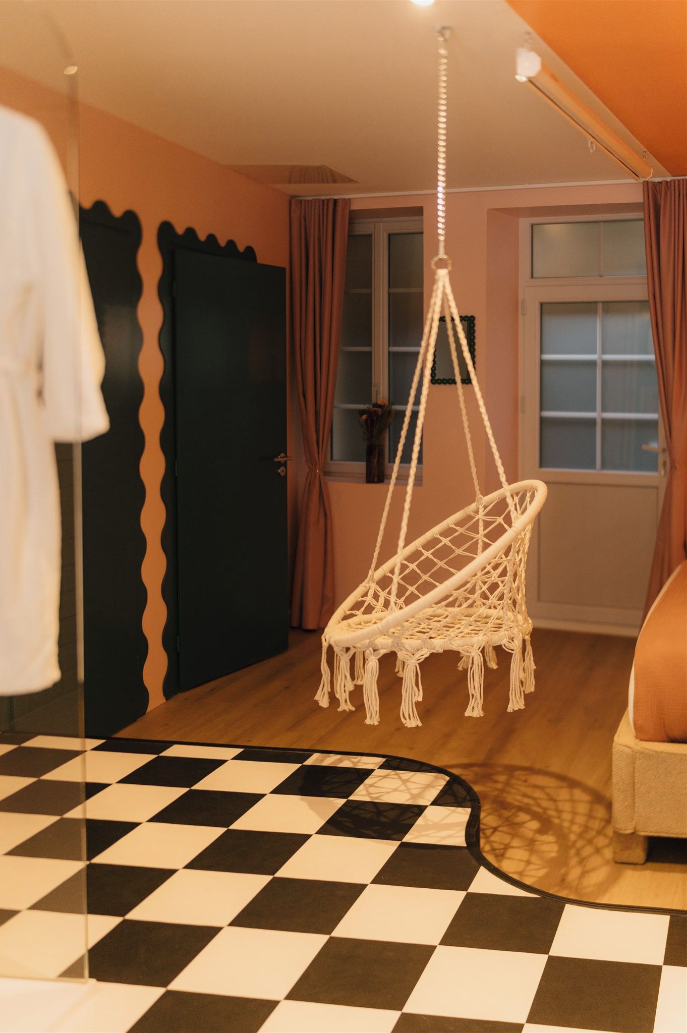 Une pièce avec un sol à carreaux, un fauteuil suspendu en macramé, un lit et une porte au motif ondulé.